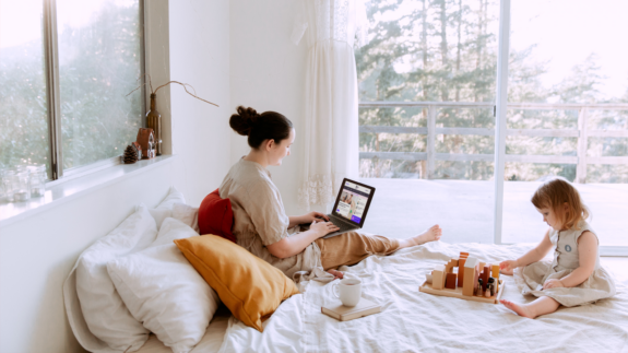 A woman looks at the All About the Mom website on a laptop while sitting on a bed with a girl who is playing with blocks.