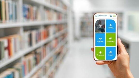 Someone holds their smartphone up with reading the J3 Foundation website in front of bookshelves in a library.